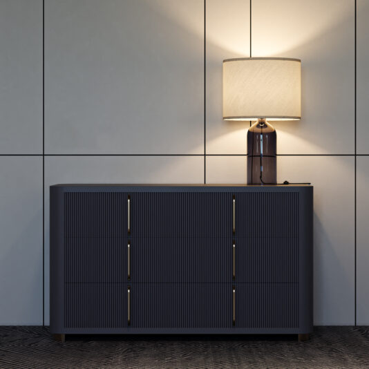 The Contemporary Ribbed Lacquered Chest of Drawers in dark gray is set against a light paneled wall, topped with a ceramic lamp and beige shade that casts a warm glow, while the floor displays a subtle geometric pattern.