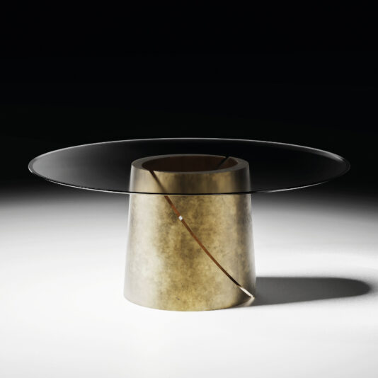 The Contemporary Ebony Veneered Dining Table (Copy) features a wide round glass top and a cylindrical metallic gold base with diagonal groove and white accent, adding modern elegance to the minimalist space highlighted by dramatic lighting.