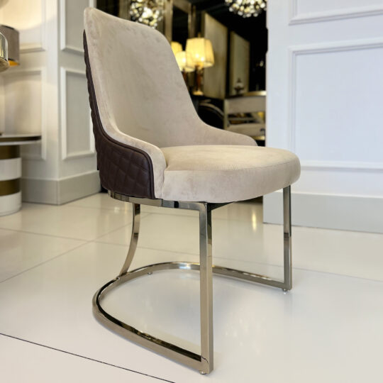 The Contemporary Two-Tone Leather Dining Chair features a beige cushioned seat and backrest, brown quilted sides, and a curved chrome base, shown on a white tiled floor in a bright, stylish interior.