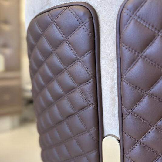 Close-up of the Contemporary Two-Tone Leather Dining Chair, featuring quilted brown leather panels with visible stitching, a slim gap between them, and a metallic accent at the base, set against a blurred background.