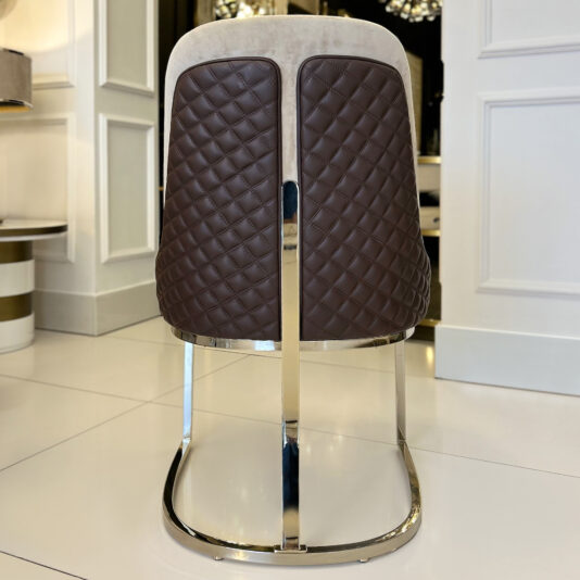 The Contemporary Two-Tone Leather Dining Chair, featuring a quilted dark brown backrest and chrome metal legs, is placed on a glossy white tiled floor in an elegant, modern room.
