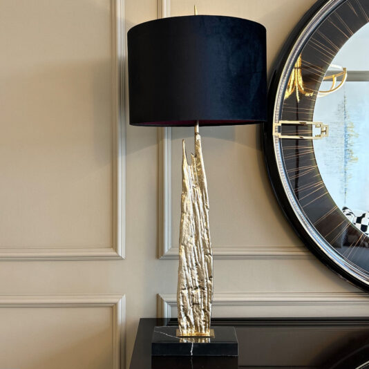 The Large Abstract Table Lamp With Velvet Shade, featuring a sculptural gold base, sits on a dark surface next to a round mirror with gold accents against a beige paneled wall.