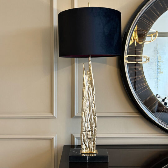 The Large Abstract Table Lamp With Velvet Shade, featuring a sculptural gold base, sits on a dark surface next to a round mirror with gold accents against a beige paneled wall.