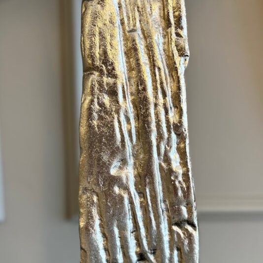 Close-up of the Large Abstract Table Lamp With Velvet Shade, featuring a vertical metallic sculpture with rough texture and shiny gold or brass finish against a softly blurred, neutral background.