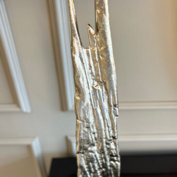 The Large Abstract Table Lamp With Velvet Shade features a vertical, textured silver metallic sculpture and is set against a light-colored wall with decorative molding.