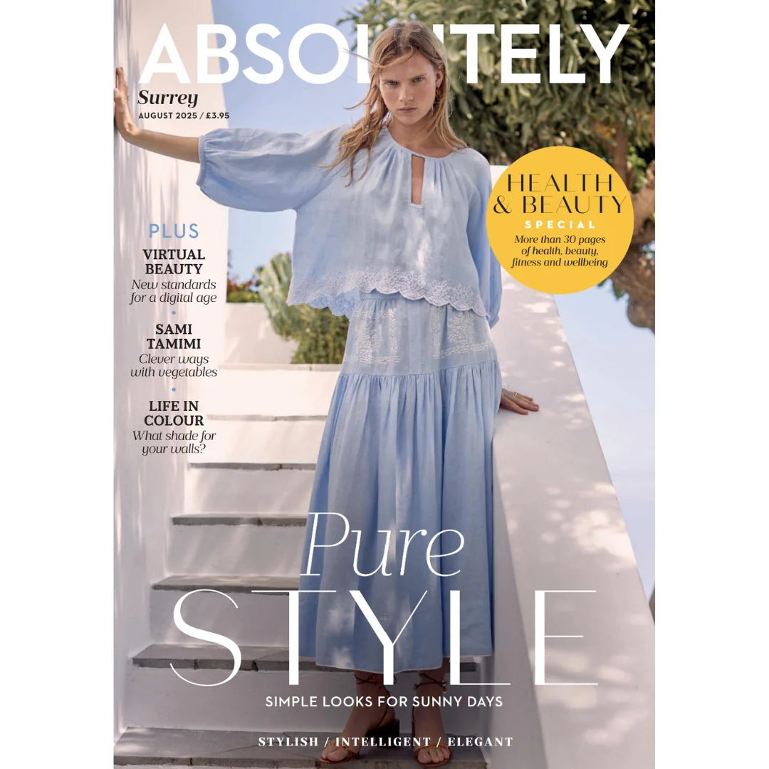 A woman wearing a light blue, flowy skirt and top poses on white steps against a bright, sunlit background. The Absolutely Surrey magazine cover features headlines about style, health & beauty, and summer fashion.