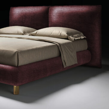 Contemporary Italian Bed The Contemporary Italian Bed features a tall, textured burgundy headboard and frame, beige sheets, two pillows, and gold-coloured legs, set against a dark background.