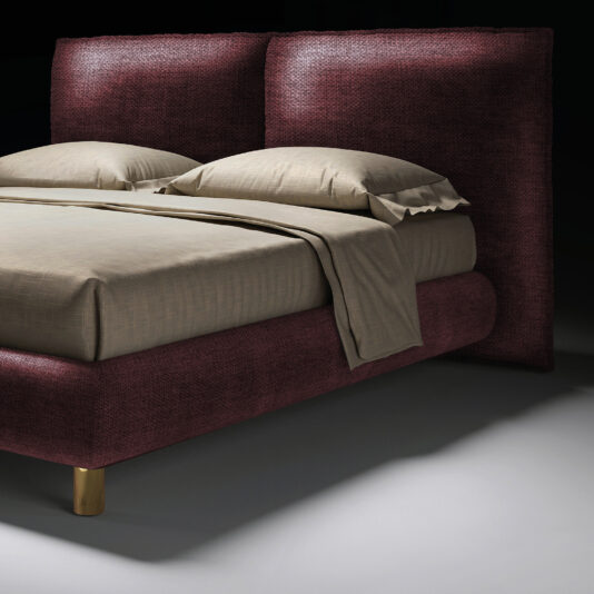 The Contemporary Italian Bed features a tall, textured burgundy headboard and frame, beige sheets, two pillows, and gold-coloured legs, set against a dark background.