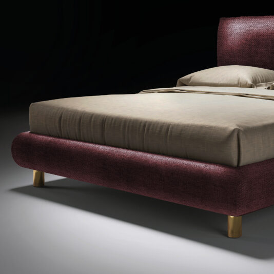 The Contemporary Italian Bed features a burgundy upholstered frame, gold legs, and beige bedding, set against a dark backdrop.