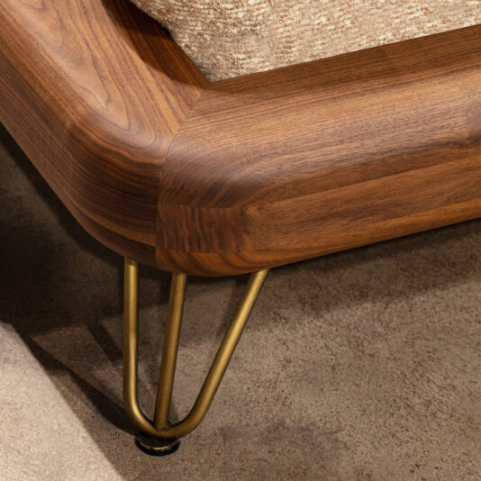 High End Contemporary Italian Bed Close-up of the High-End Contemporary Italian Bed corner, featuring rounded wooden edges, gold hairpin metal legs, and a textured beige cushion, placed on a brown carpeted floor.