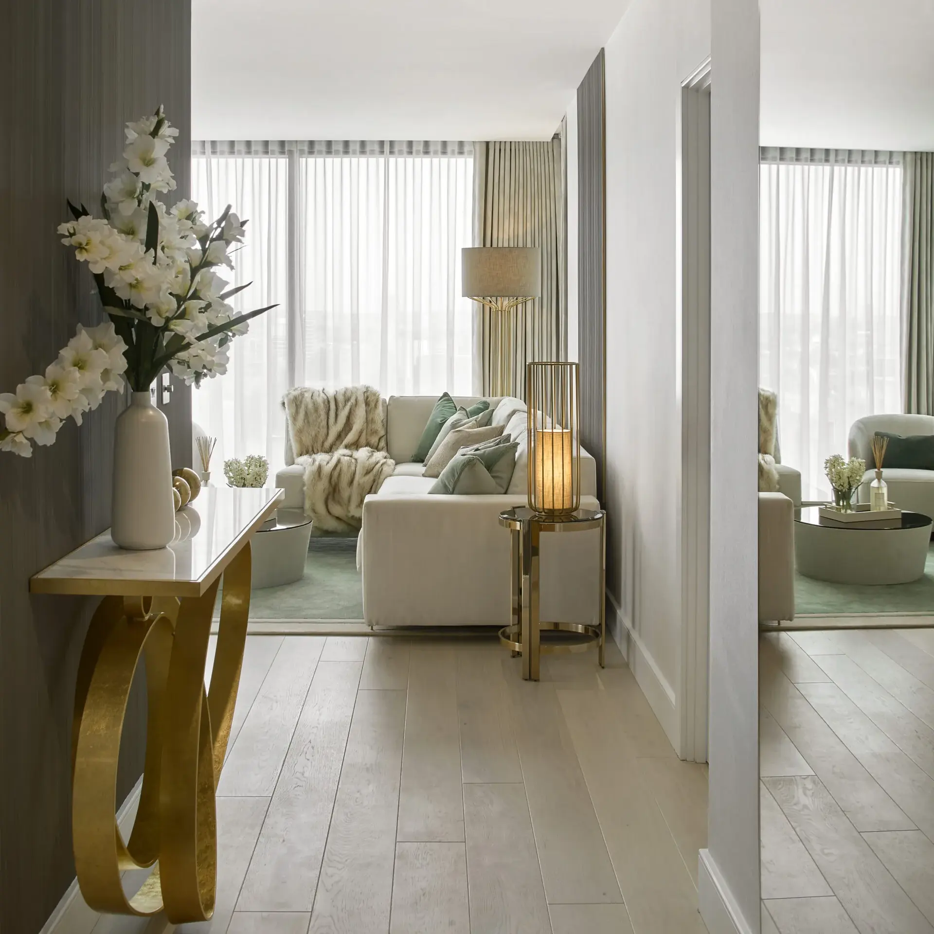 A modern living room view from a hallway. The space features a white sofa, green accents, and a floor lamp. A console table with white flowers and a large mirror are in the foreground. Light wood flooring complements the neutral and elegant decor.