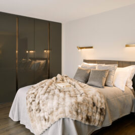 Modern bedroom with a double bed, grey pillows, a faux fur blanket, two bedside tables with books, and built-in glossy wardrobes. Warm wall-mounted lights illuminate the neutral-toned, cosy space.
