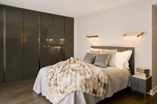 Modern bedroom with a double bed, grey pillows, a faux fur blanket, two bedside tables with books, and built-in glossy wardrobes. Warm wall-mounted lights illuminate the neutral-toned, cosy space.