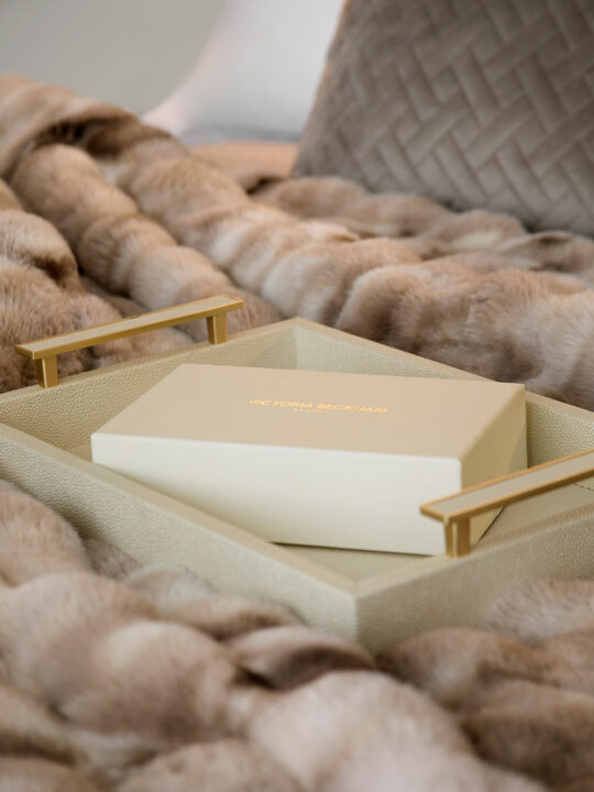 A cream-coloured box with gold lettering reading "Victoria Beckham" sits on a beige tray with gold handles, placed on a soft, light brown faux fur blanket.