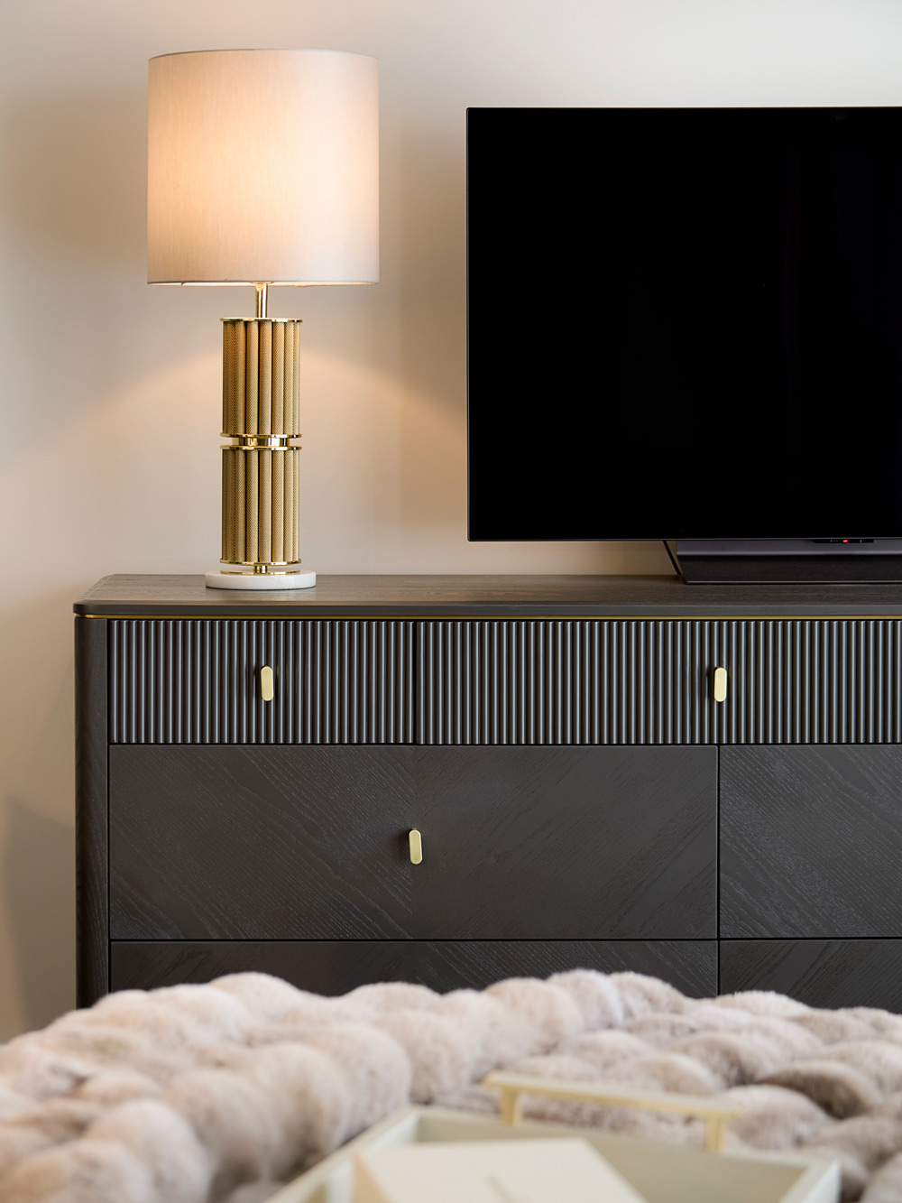 A modern bedroom features a dark chest of drawers with gold handles, a gold and white table lamp, and a flat-screen television. A soft, light-coloured blanket is draped over the foot of the bed in the foreground.