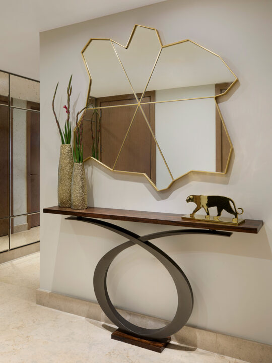 A modern entrance hall features a sculptural console table with a curved black metal base, two tall textured vases with flowers, a decorative gold animal figurine, and a large abstract gold-framed mirror on the wall.