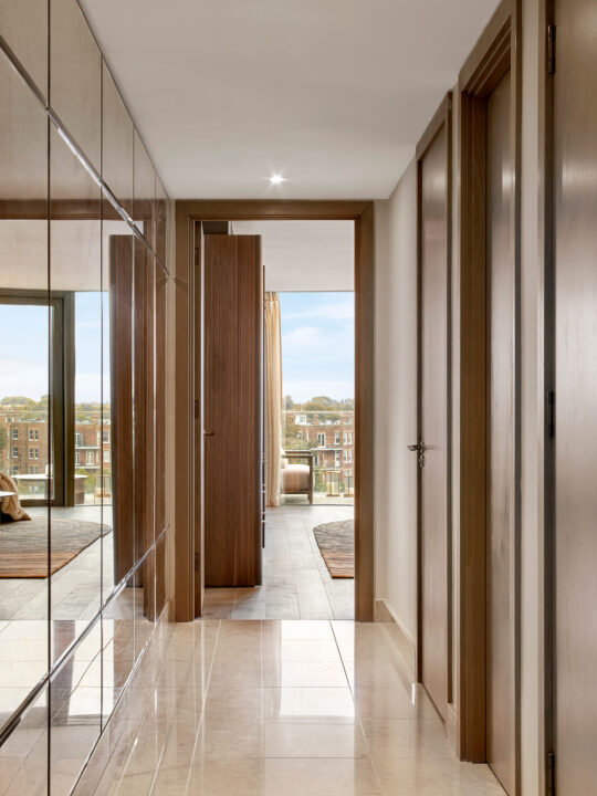 A modern hallway with beige tiled floors, wooden doors, and mirrored wardrobes leads to a bright room with large windows overlooking buildings and trees outside.
