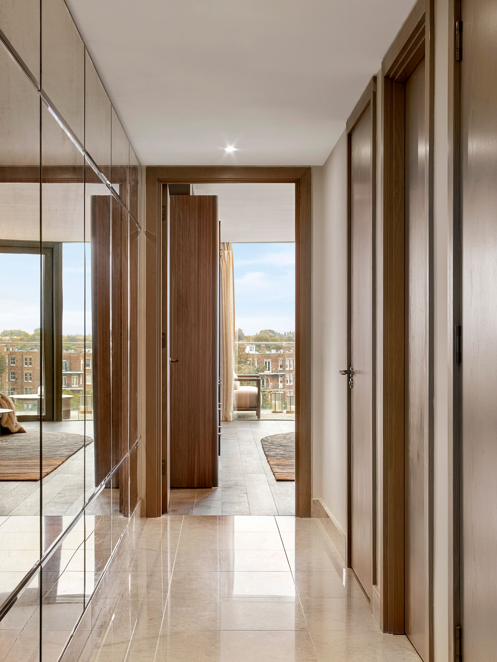 A modern hallway with beige tiled floors, wooden doors, and mirrored wardrobes leads to a bright room with large windows overlooking buildings and trees outside.