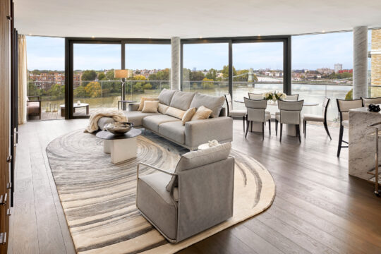 A modern, spacious living and dining area with large floor-to-ceiling windows, neutral-toned furniture, a round rug, and a view of a river and cityscape outside.