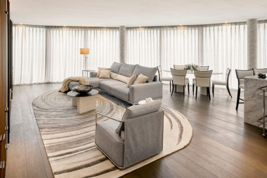 A modern living and dining room with floor-to-ceiling sheer curtains, light grey furniture, a round patterned rug, a marble coffee table, a dining table with six chairs, and wooden floors.