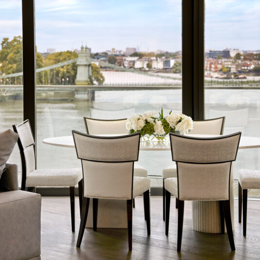 A modern dining area with six upholstered chairs around a round white table holding a flower arrangement, set beside large windows overlooking a river and cityscape.
