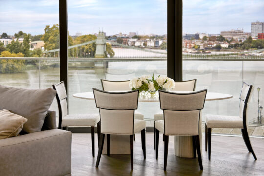 A modern dining area with six upholstered chairs around a round white table holding a flower arrangement, set beside large windows overlooking a river and cityscape.