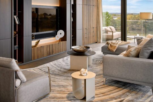 Modern living room with neutral-toned furniture, a patterned rug, a large TV on a wooden wall unit, decorative objects on tables, floor-to-ceiling windows, and a view of greenery outside.