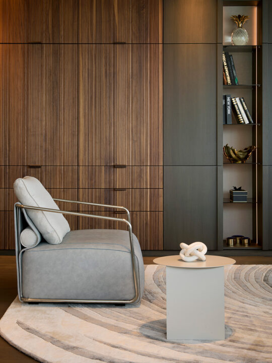A modern living room with a grey armchair, a round side table with a white decorative object, a curved patterned rug, wooden panelled walls, and a built-in shelf with books and decorative items.
