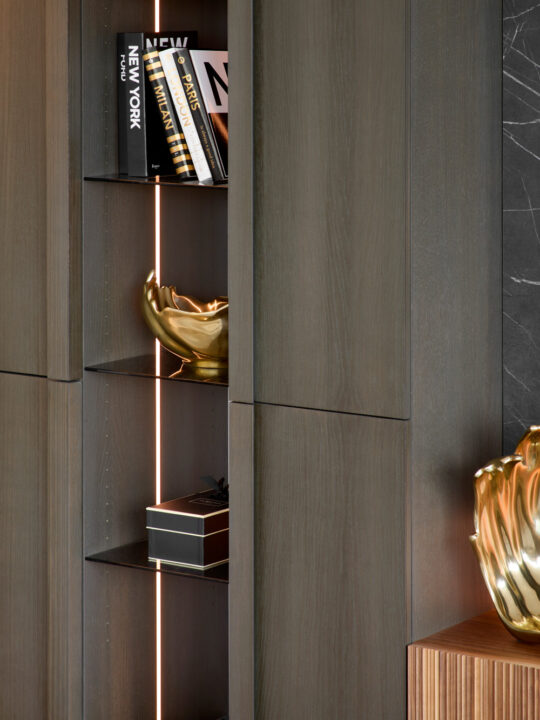 A modern wooden shelving unit with books, a decorative gold bowl, a small black gift box with a white ribbon, and warm vertical lighting. Part of a marble wall and wooden surface are also visible.