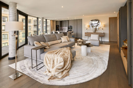 A modern, spacious living room with floor-to-ceiling windows, a grey corner sofa, a round rug, fur throw, and neutral décor. Open kitchen and dining area are visible in the background with stylish furnishings.