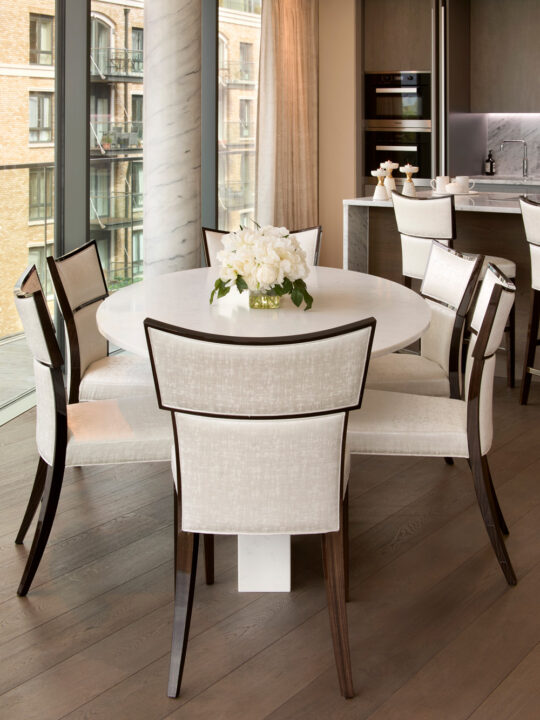 A modern dining area with a round white table surrounded by six upholstered chairs, featuring dark wooden frames, on a light wood floor. A vase of white flowers sits at the centre of the table. Large windows and a kitchen are in the background.