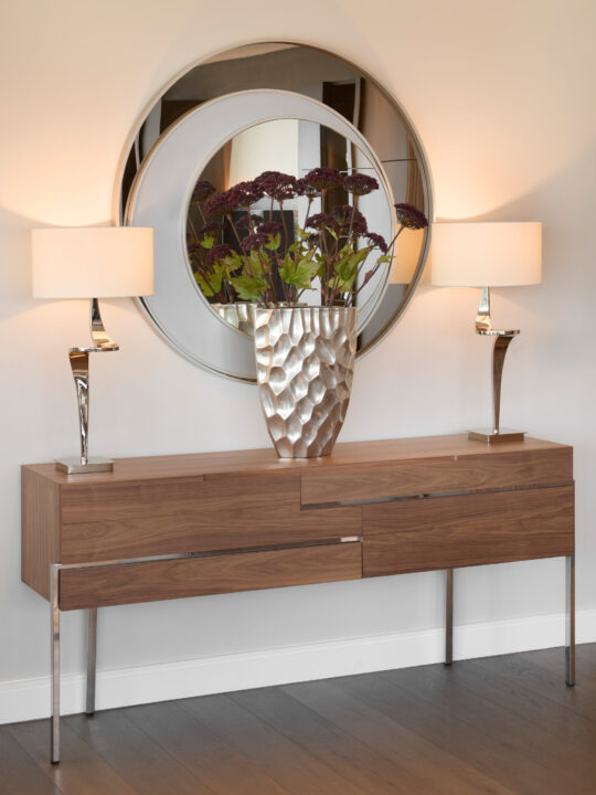 A modern wooden console table with metal legs holds a textured silver vase of dark red flowers, flanked by two sculptural lamps. A large round mirror is mounted on the wall behind, reflecting the arrangement.