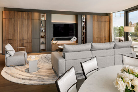 Modern living room with a large grey corner sofa, armchair, round rug, wall-mounted TV, built-in wooden cupboards, and a round dining table with white chairs and flowers, next to large windows with city views.