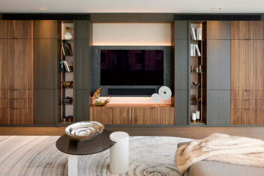 Modern living room with built-in wooden cupboards and shelves, a large flat-screen TV, decorative sculptures, books, and vases. A round coffee table with a bowl and a cosy beige throw is in the foreground.