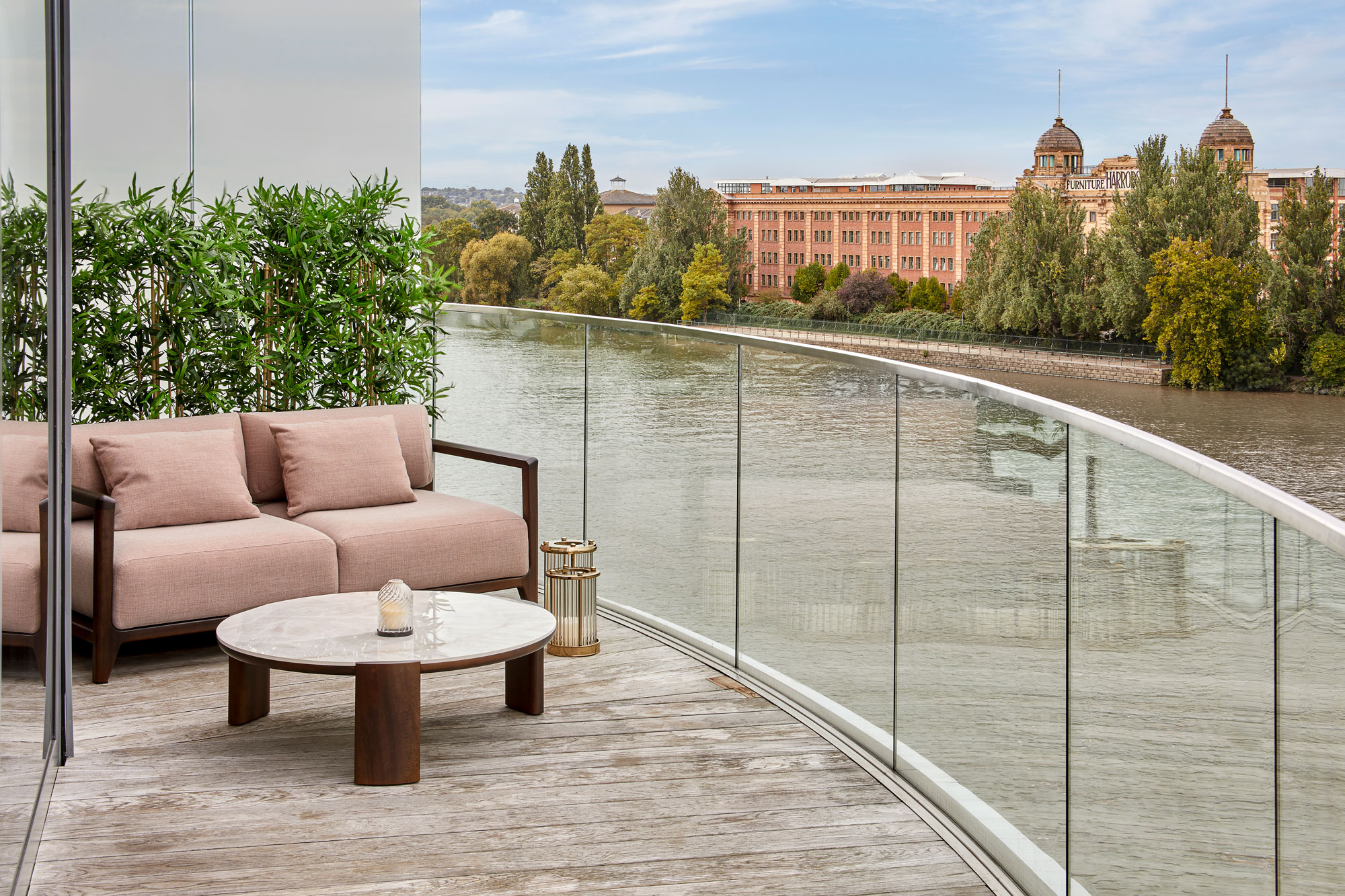 A modern balcony with a glass balustrade overlooks a river and historic brick building. The balcony has a pink cushioned sofa, a round marble coffee table, a candle, a lantern, and potted greenery.