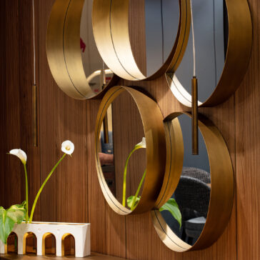 Modern Floating Wall Mirror Cluster A modern interior showcases the Modern Floating Wall Mirror Cluster with circular gold frames on a wood-paneled wall, accompanied by a white vase with three calla lilies and a small decorative white piece on the wooden surface.