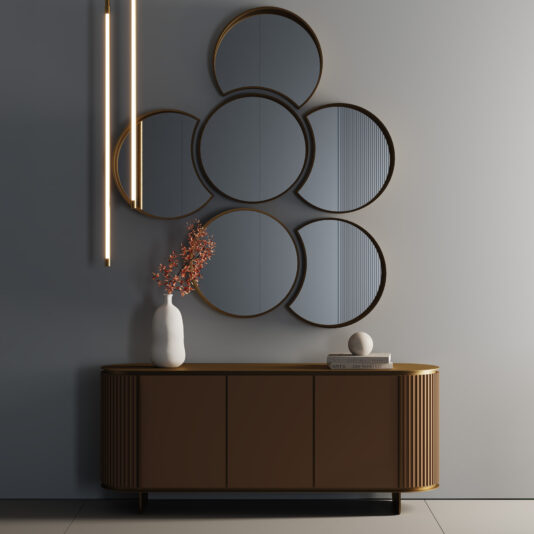A modern entrance features a brown sideboard, a white vase with orange branches, stacked books, decorative stone, and the Modern Floating Wall Mirror Cluster of six round mirrors above, with vertical pendant lights on the left.