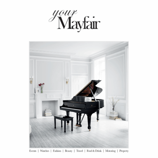A grand piano with a matching stool sits in an elegant, light-filled room with white panelled walls, large windows, and minimal decor. The image is featured on the cover of "your Mayfair" magazine.
