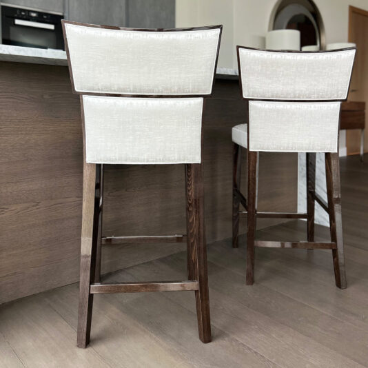 Two Classic Deco Style Bar Stools with dark wooden legs and light upholstery are placed at a wood-panelled kitchen island in a contemporary home.