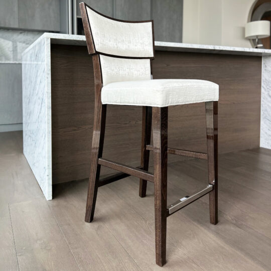 The Classic Deco Style Bar Stool features a modern wooden frame and white cushioned seat and backrest, shown on a light wood floor in front of a marble-topped kitchen island.