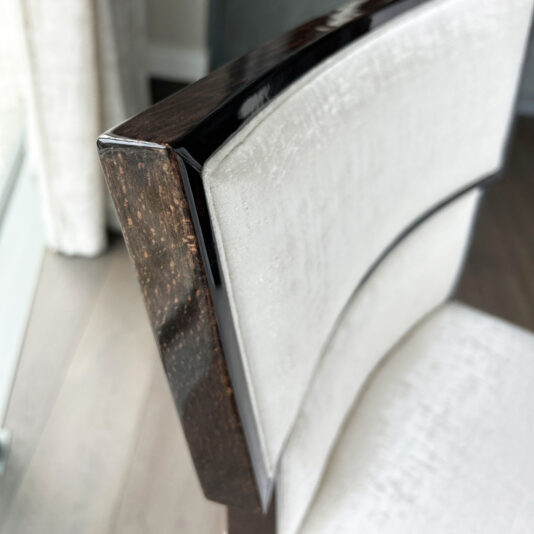 Close-up of the Classic Deco Style Bar Stool, featuring a dark wood frame and light upholstered backrest, positioned on a wooden floor near a window with net curtains.