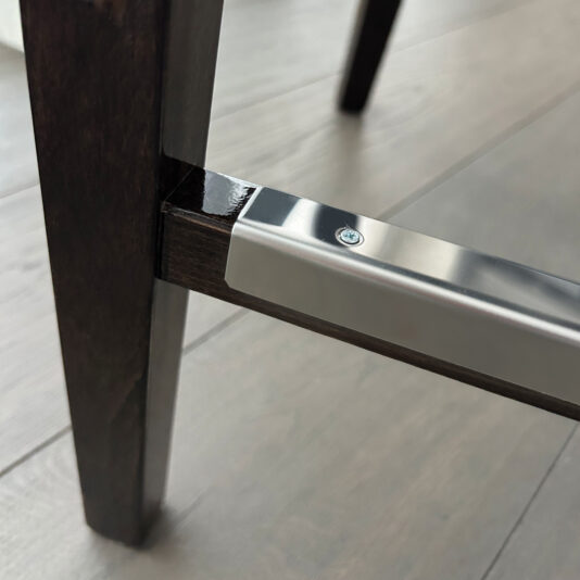 Close-up of the Classic Deco Style Bar Stool’s wooden leg with a polished metal bracket and screw, shown resting on a light-toned wood floor.