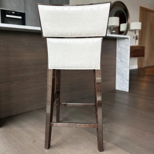 The Classic Deco Style Bar Stool features a tall wooden frame and light beige upholstered backrest and seat, shown on a light wood floor in a modern kitchen with brown cupboards and a marble worktop.