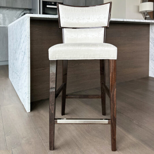 The Classic Deco Style Bar Stool features a white cushioned seat and backrest, dark wooden legs, and a metal footrest, positioned on a light wood floor in front of a marble kitchen island.