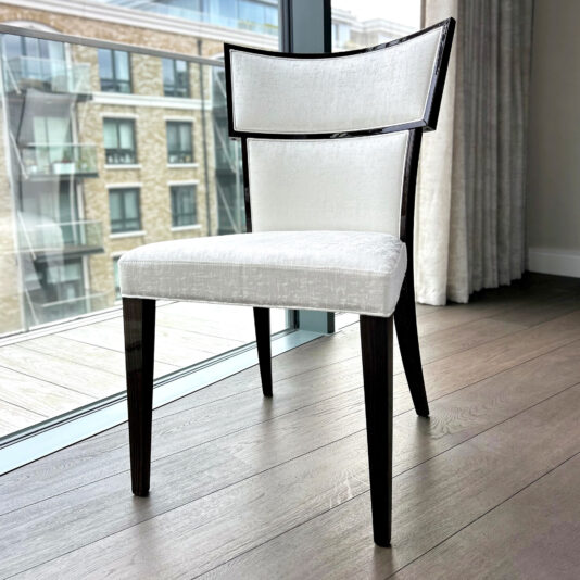 The Classic Deco Style Dining Chair, with dark wooden legs and frame and a white upholstered seat and backrest, stands on a wooden floor near a large window overlooking city buildings.
