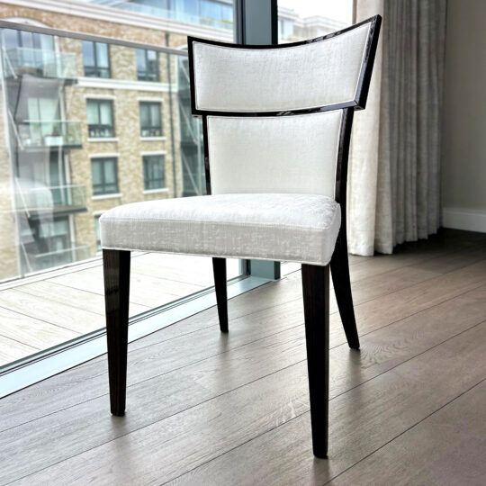 The Classic Deco Style Dining Chair, with dark wooden legs and frame and a white upholstered seat and backrest, stands on a wooden floor near a large window overlooking city buildings.