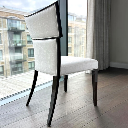 The Classic Deco Style Dining Chair, featuring a white upholstered seat and backrest with a dark wooden frame and legs, sits by a large glass window that overlooks a balcony and nearby brick block of flats.