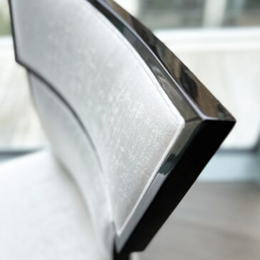Classic Deco Style Dining Chair A Classic Deco Style Dining Chair features a glossy black wood frame and a light grey upholstered backrest, placed near a window with blurred outdoor scenery in the background.