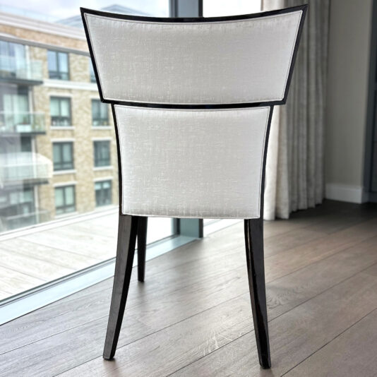 The Classic Deco Style Dining Chair features light upholstery and dark legs, set on wooden flooring by large windows with views of a block of flats.