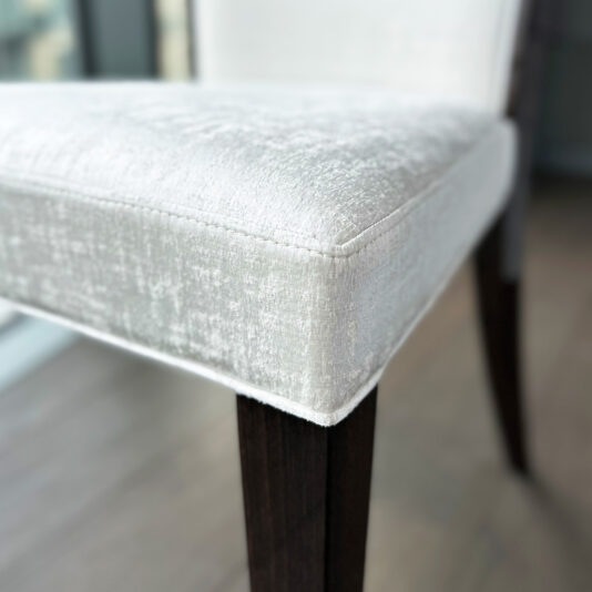 Close-up of the Classic Deco Style Dining Chair featuring a textured, light upholstery on the seat and backrest, dark wooden legs, set on a wooden floor with a softly blurred background.